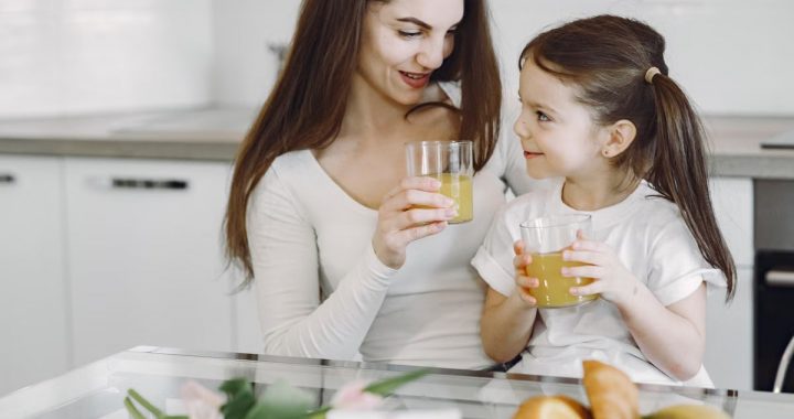 Happy mother and daughter in casual clothes drinking juice and looking at each other while having breakfast together in contemporary kitchen