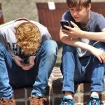 Two teenagers seated outdoors, focused on their smartphones, embracing modern technology.