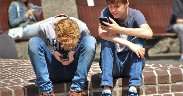 Two teenagers seated outdoors, focused on their smartphones, embracing modern technology.