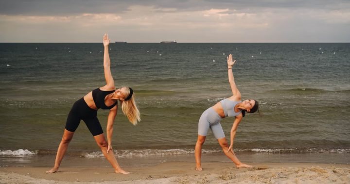 Two women exercising at a beach, performing yoga poses with the ocean in the background during sunset.